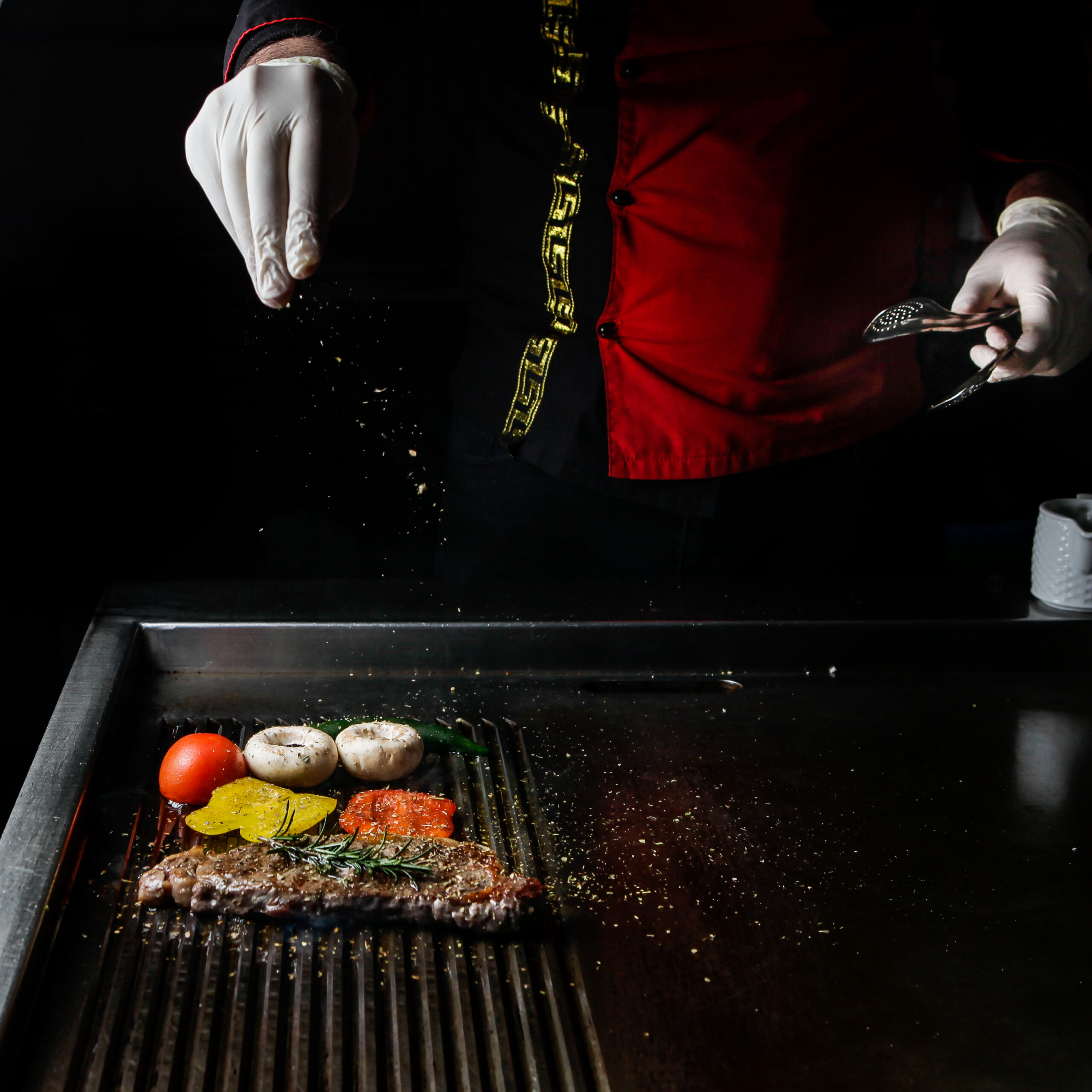 side view grilled steak with rosemary and tomatoes and human hands in barbecue on dark background